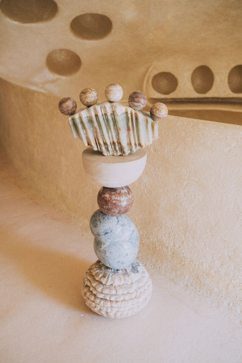A sculptural arrangement of stacked, round and textured stones and ceramics stands on a beige floor against a curved, sandy-colored wall with circular indentations