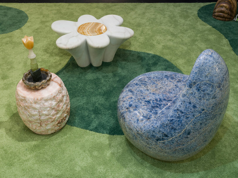 A round blue stone chair by Sten Studio, a pink stone side table with decor, and a white flower-shaped coffee table rest elegantly on a green patterned carpet.