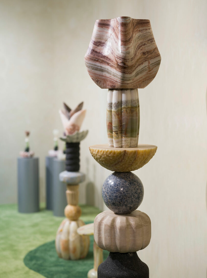 A close-up of a colorful, abstract stone sculpture by Sten Studio, composed of stacked geometric shapes, with more similar sculptures displayed in the background on cylindrical stands.