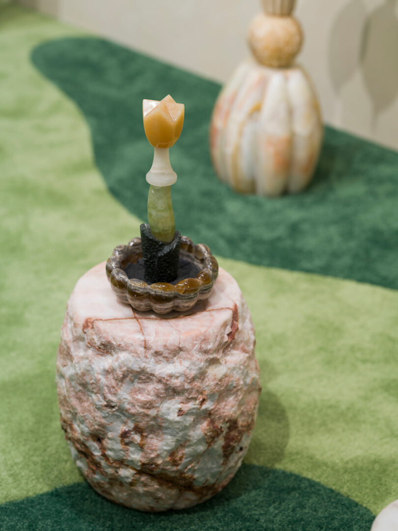 A small sculpture by Sten Studio, crafted from stone and minerals and shaped like a flower, sits on a textured stone pedestal with green carpet and another sculpture in the background.