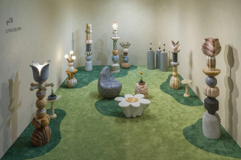 Art installation by Sten Studio in a gallery room featuring stone sculptures shaped like abstract flowers and plants on green carpet with organic patterns. The walls are plain and neutral-colored.