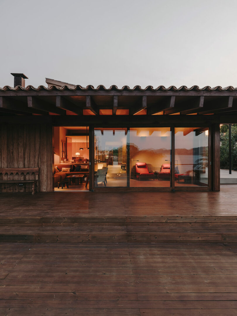 Exterior view of a modern wooden house at dusk with large glass doors showing a warmly lit interior and outdoor reflections on the windows.
