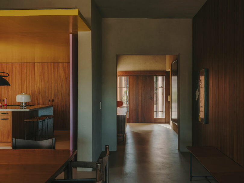 Modern interior with wood-paneled walls, open kitchen area, dining table, and illuminated hallway leading to a front door with vertical slats.