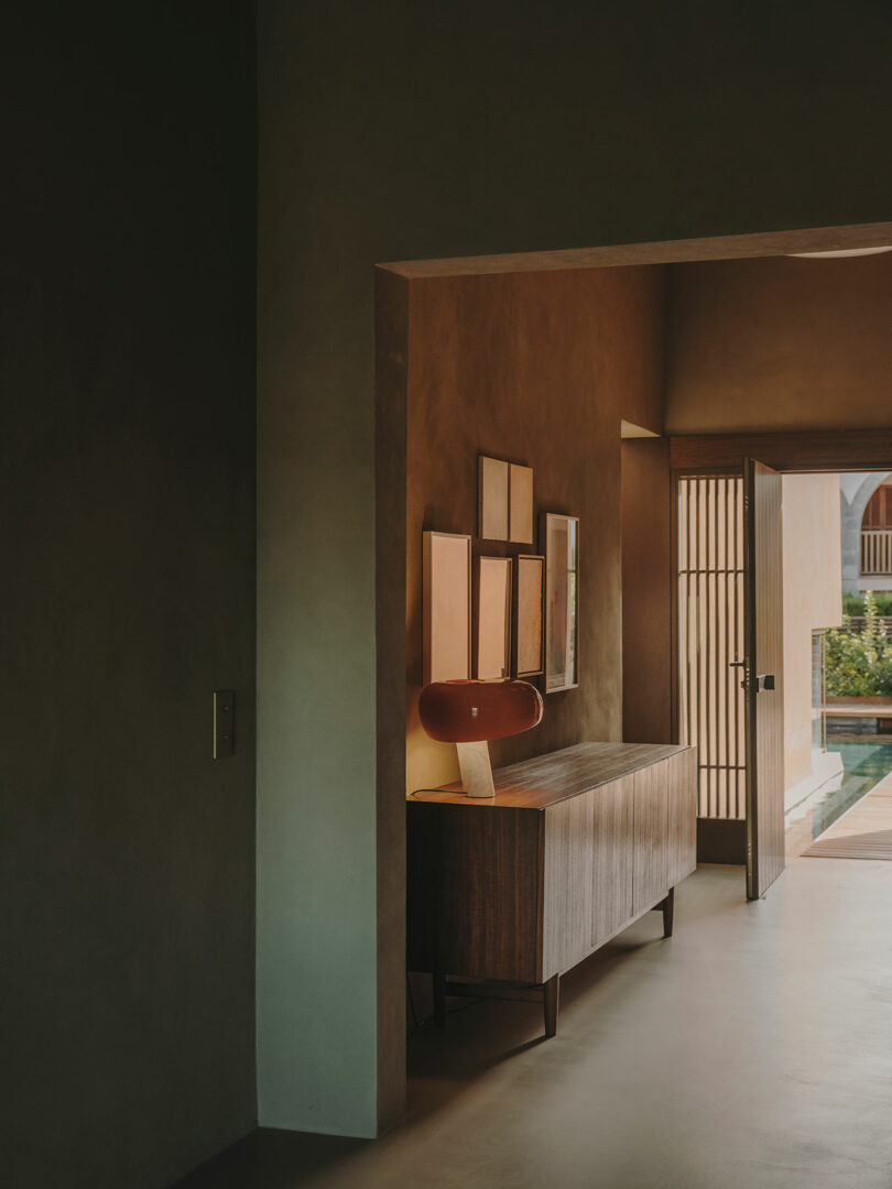 A narrow hallway with muted walls features a wooden sideboard, a red lamp, five framed pictures, and an open door leading to a sunlit outdoor area with greenery.
