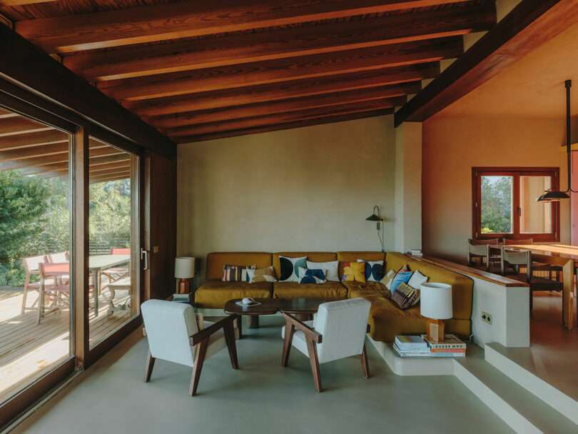 Modern living room with exposed wooden beams, mustard sectional sofa with pillows, two white armchairs, a round coffee table, and large windows opening to a deck with outdoor seating.