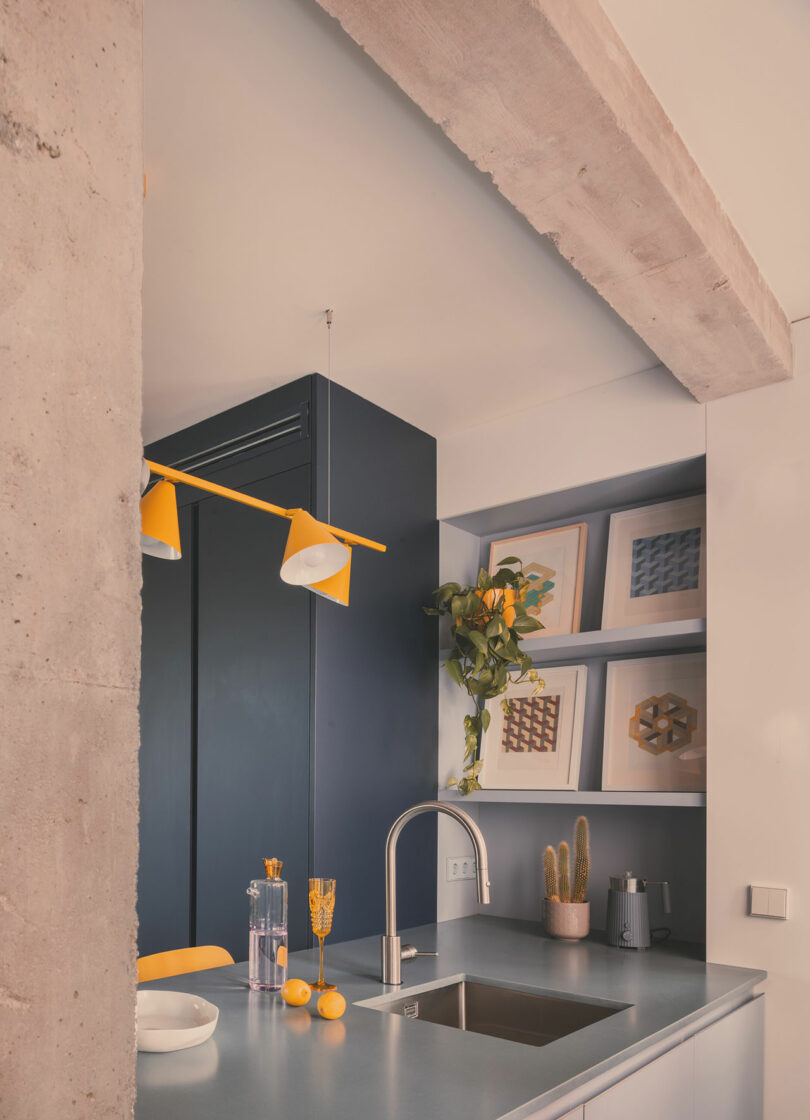 Modern kitchen with a gray countertop, built-in sink, yellow accents, wall art, potted plants, and a concrete beam overhead.