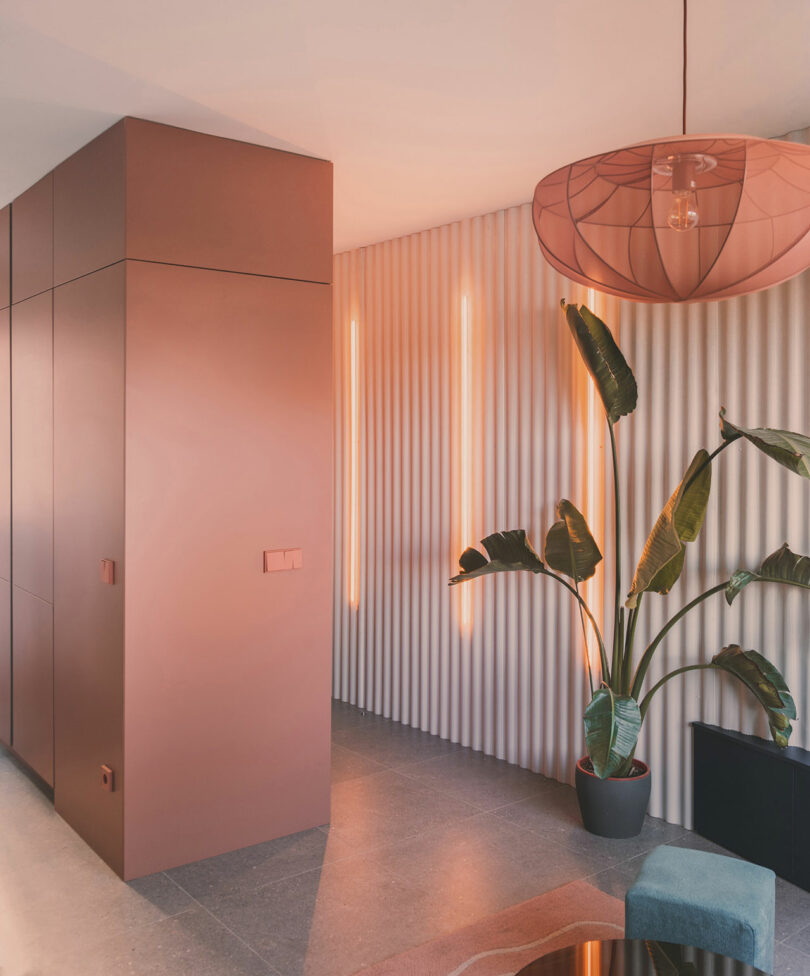 Modern interior hallway with pink built-in cabinets, ribbed beige wall paneling, vertical orange lights, a large potted plant, and a pink pendant lamp.