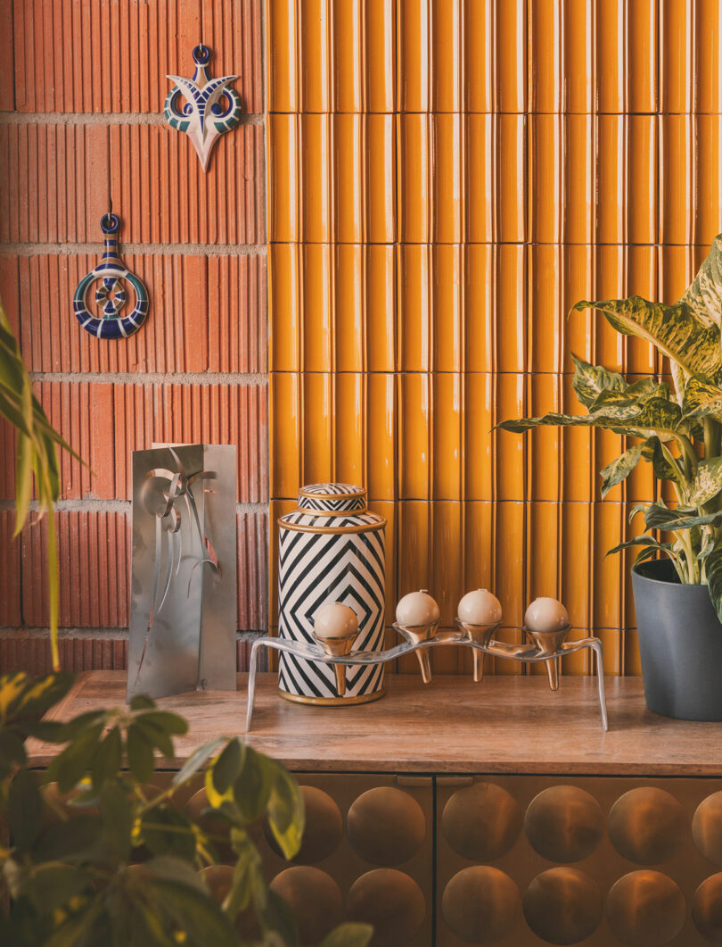 A decorative tabletop with a geometric jar, four egg holders, potted plants, and wall art against a yellow tiled and red brick background.