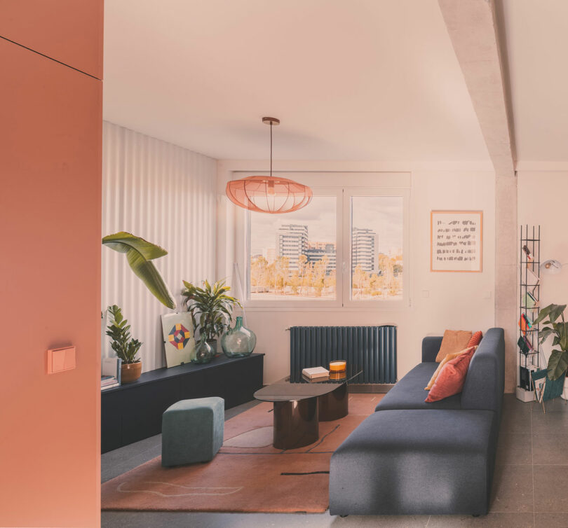 A modern living room with a blue sofa, coffee table, plants, a geometric rug, and large window showing city buildings outside.