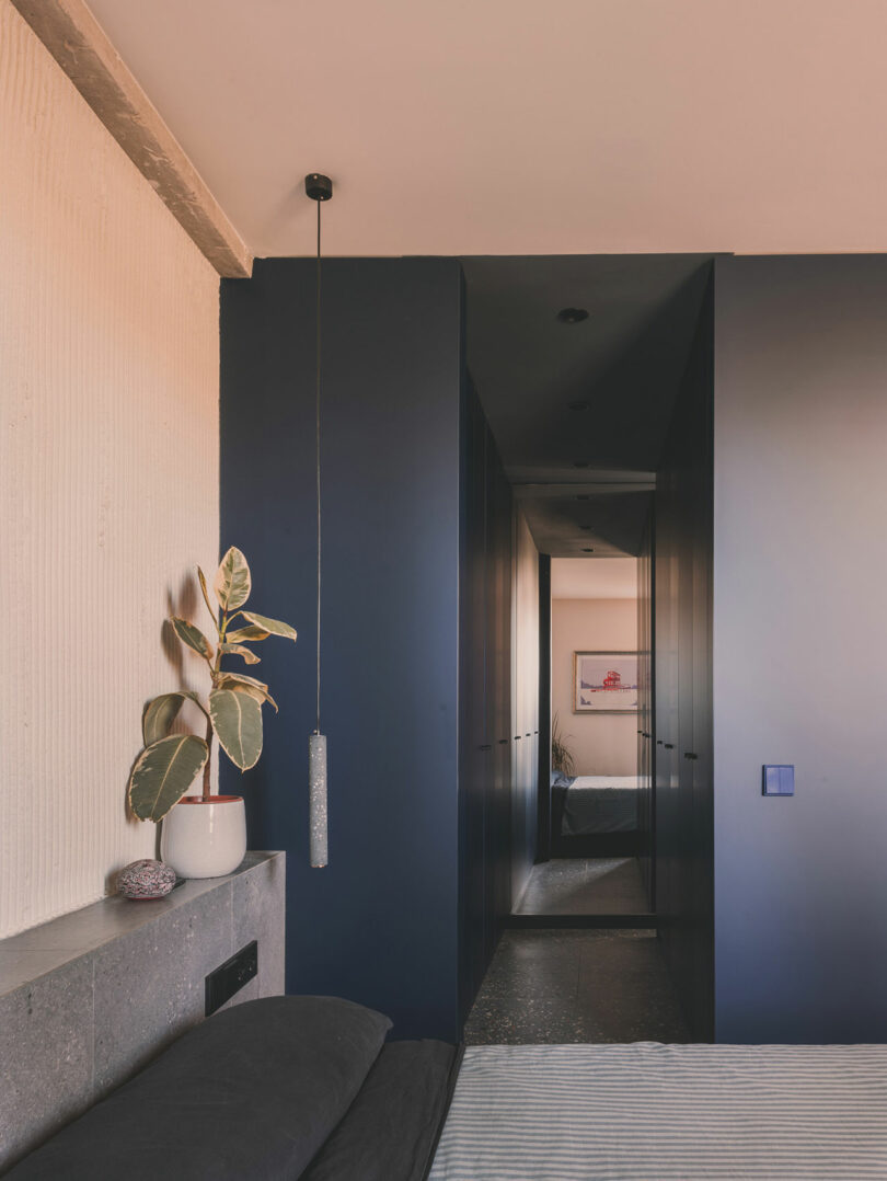 A modern bedroom features a potted plant on a concrete ledge, dark blue walls, a minimalist pendant light, and a framed artwork visible through a narrow hallway.