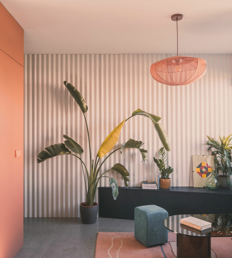 Modern living room with a corrugated white wall, large potted plants, a black console, books, a geometric art piece, and a pink pendant light.
