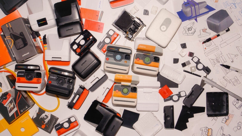 A variety of Polaroid cameras, components, packaging, and design sketches are spread out on a white table.