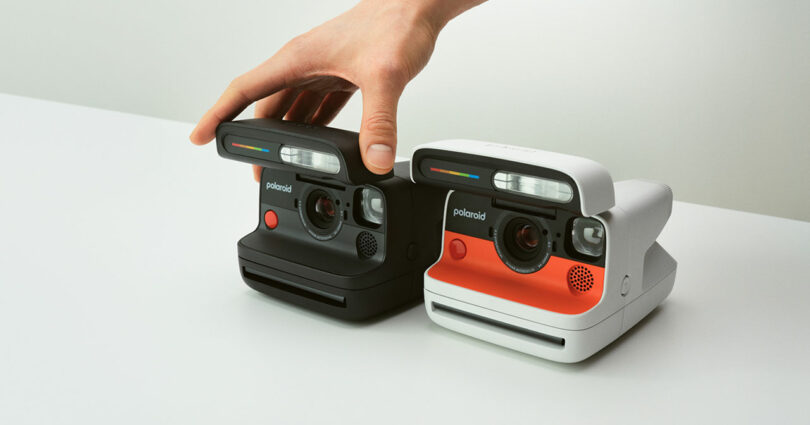 A hand touches the top of a black Polaroid instant camera next to a white and red Polaroid camera on a white surface.