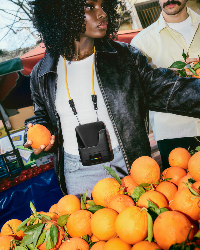 A person wearing a black leather jacket and white shirt selects oranges at an outdoor market stand; another person stands nearby.