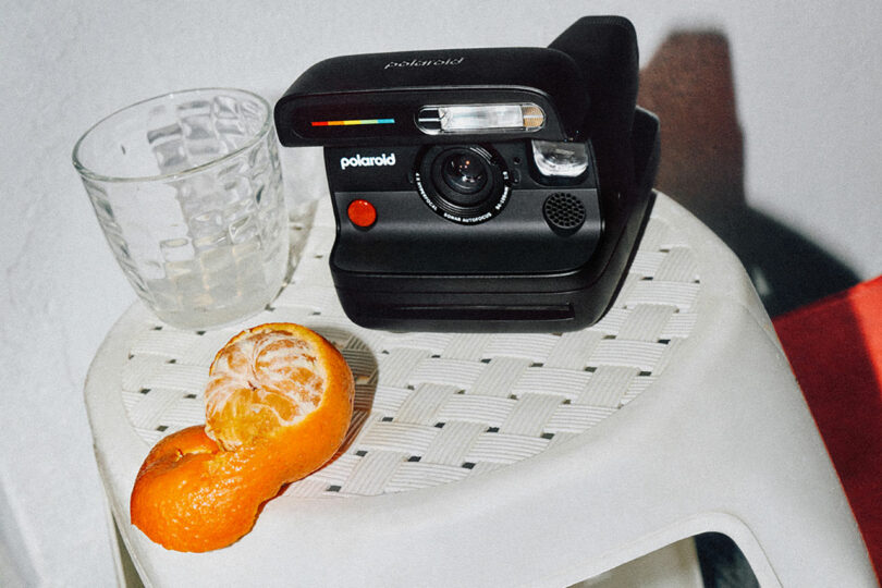 A black Polaroid instant camera, a clear glass, and a partially peeled orange sit on a white woven stool.