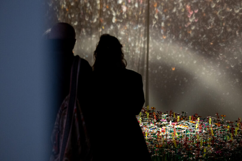 Two people stand in shadow observing a colorful, illuminated art installation featuring many small, flower-like objects with light projected on the background wall.