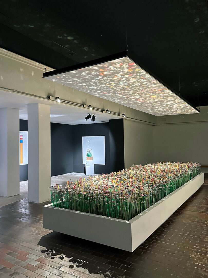 A modern art installation features a rectangular bed of colorful artificial flowers, with a large illuminated panel suspended above in a gallery space, beautifully reflecting diversiveness and mo man tai's spirit.