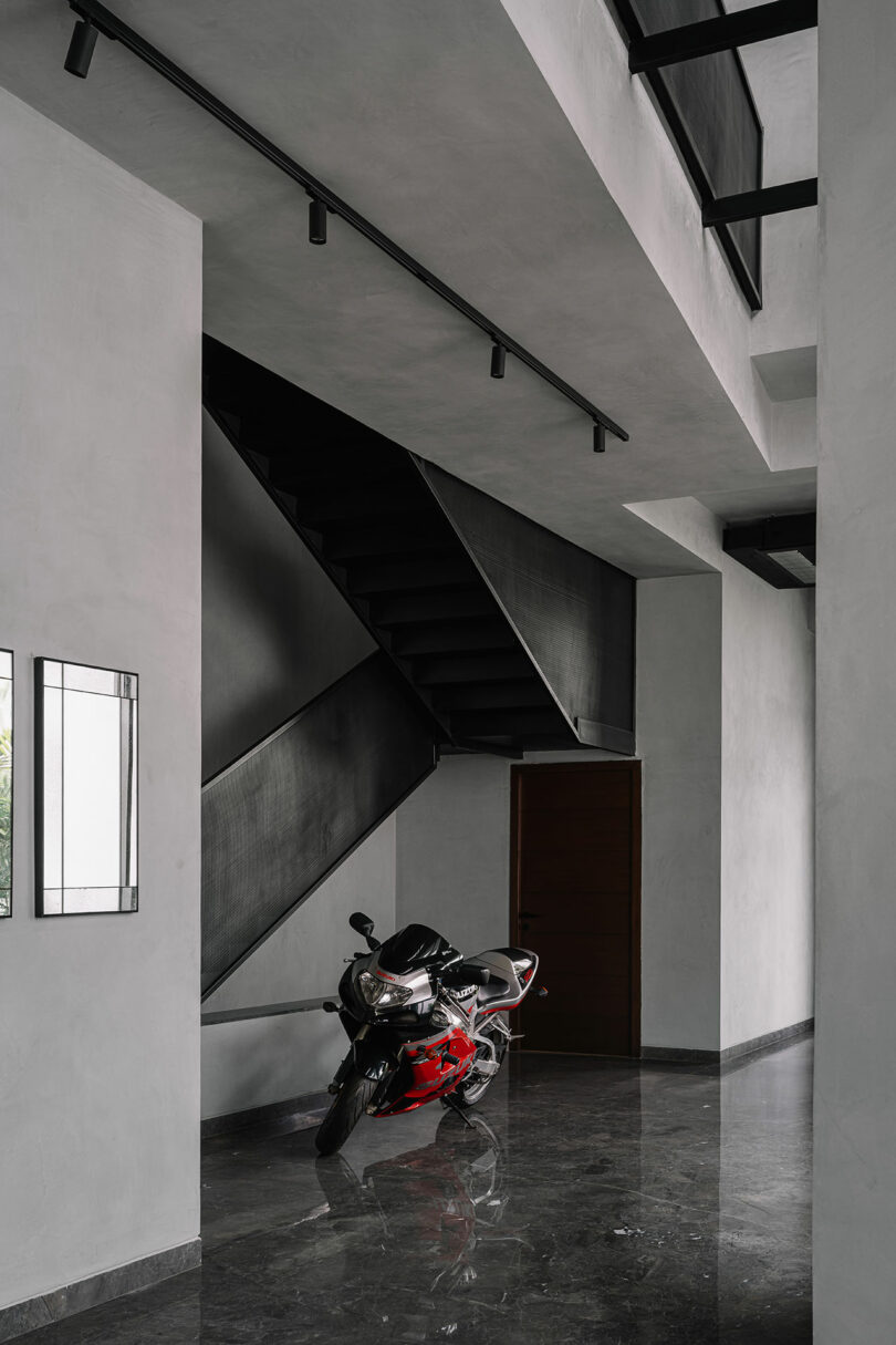 A black and red motorcycle is parked indoors on a polished dark floor beneath a modern black staircase with minimalist gray walls.