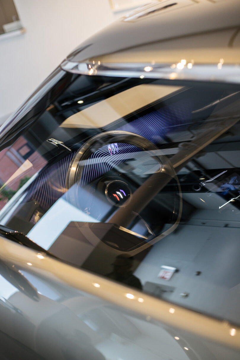 Close-up view through a car windshield showing the steering wheel and reflections of building interiors on the glass.