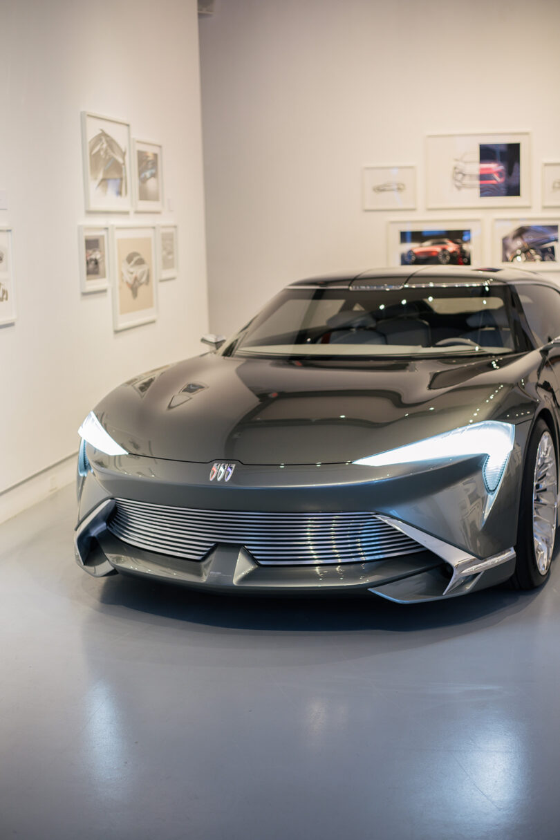 A sleek, silver Buick concept car is displayed in a modern showroom with framed automotive sketches on the walls.