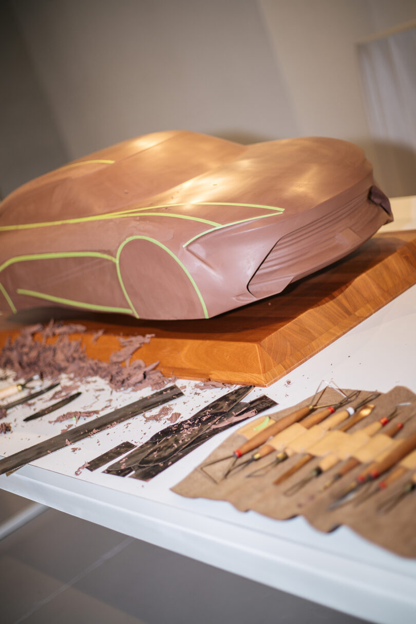 A clay model of a car sits on a wooden base, with carving tools and shavings arranged on a nearby white table.