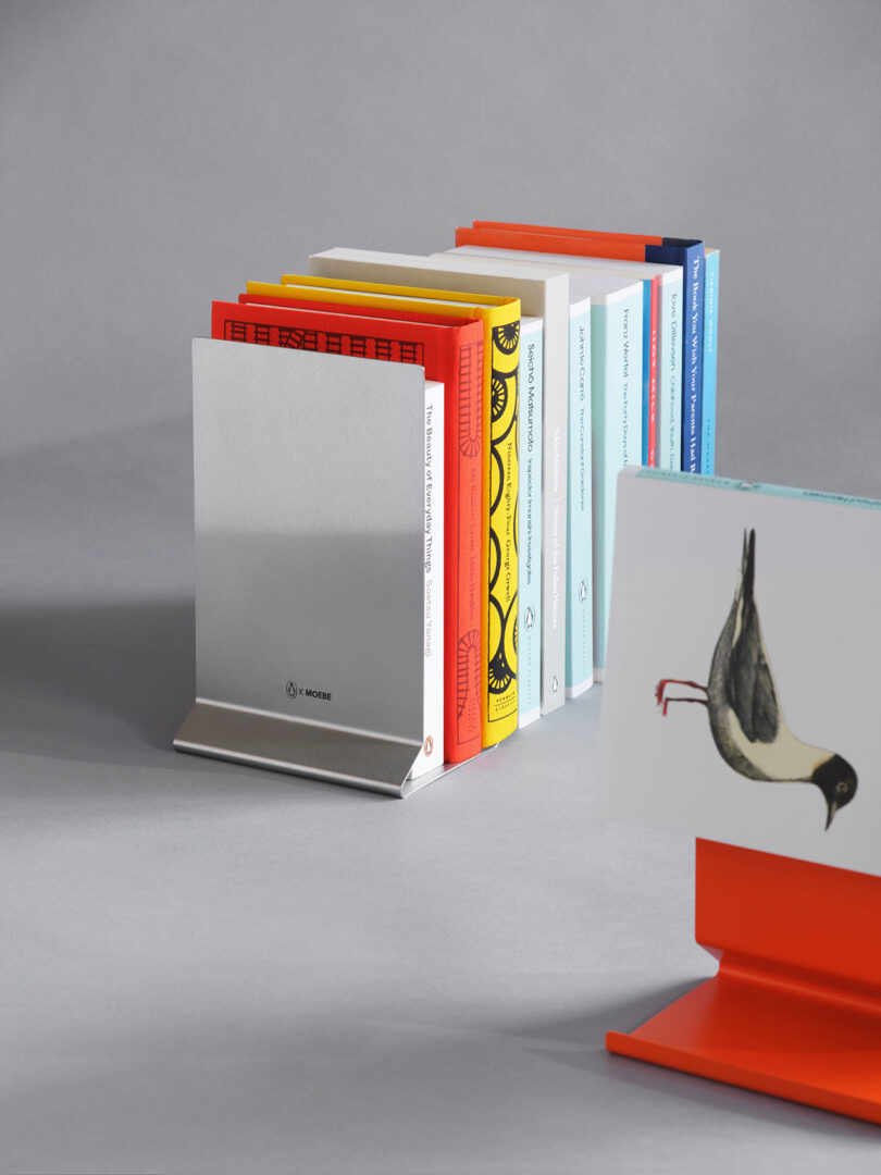 A row of colorful books is held upright by two modern metal bookends, one plain silver and one orange with a bird illustration.