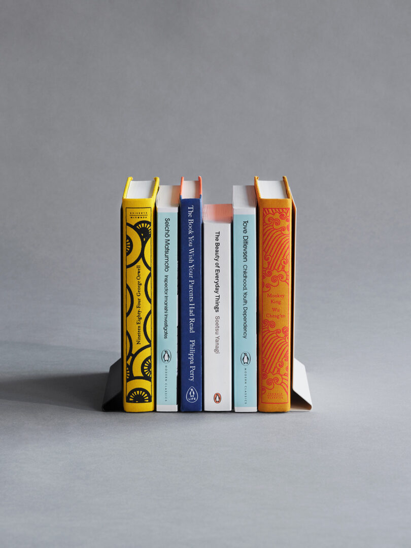 Five hardcover books of varying colors are standing upright between two gray metal bookends against a plain gray background.