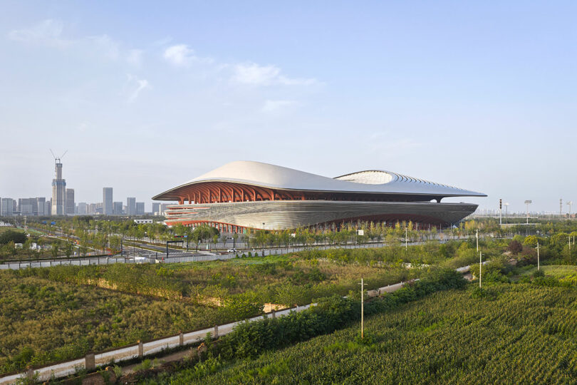 Large, modern stadium with a curved, dome-like roof surrounded by greenery and city buildings in the background under a clear sky
