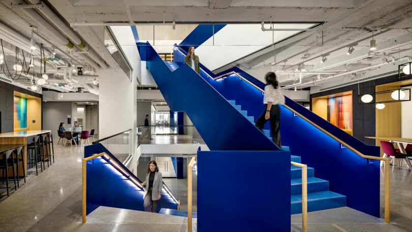 A modern office interior featuring a bold blue staircase, open workspaces, tables, and people walking throughout the area