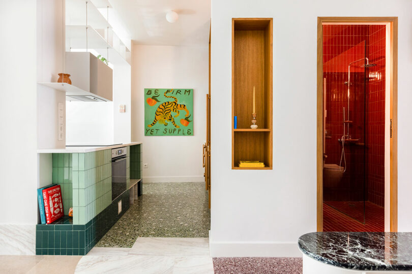 Modern kitchen with green tiles, open shelving, and a visible red-tiled shower room. A colorful 