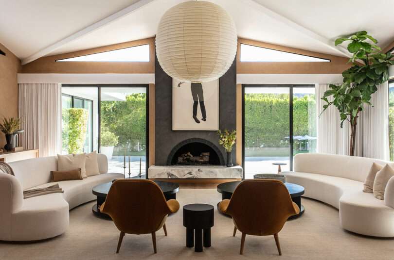 Modern living room with curved white sofas, two brown armchairs, round tables, a marble fireplace, large paper lantern, wall art, tall plant, and floor-to-ceiling windows