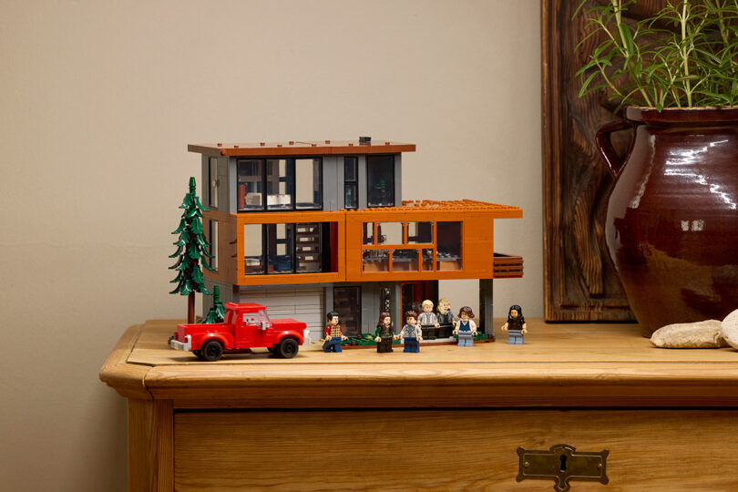 A LEGO modern house model with a red truck, six LEGO figures, and a tree displayed on a wooden chest next to a large brown pot with green plant