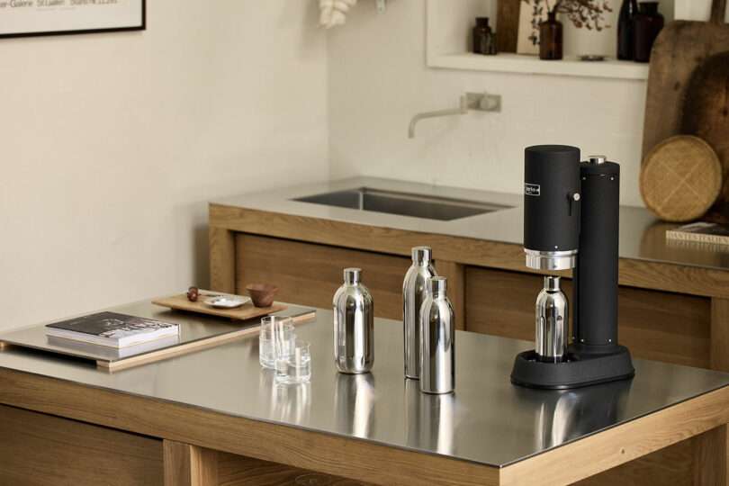 A black soda maker and three metallic bottles are placed on a metal countertop in a modern kitchen with wooden cabinets