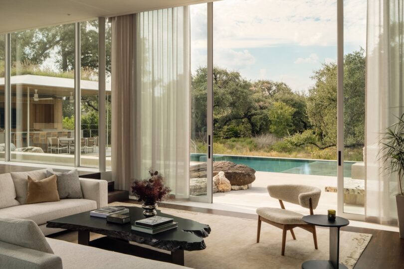 Modern living room with large floor-to-ceiling windows overlooking a swimming pool, patio, and trees; neutral-toned furniture in calming skim milk hues and natural light fill the space.