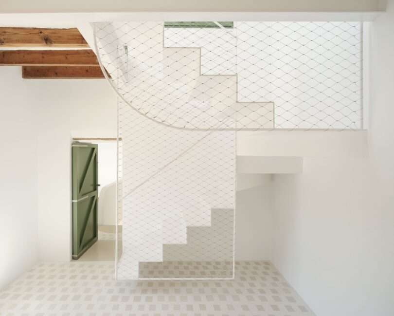 White staircase with geometric lines, a safety net barrier, green door, wooden ceiling beams, and patterned light-colored floor tiles evoke a minimalist interior reminiscent of Skim Milk design sensibilities.