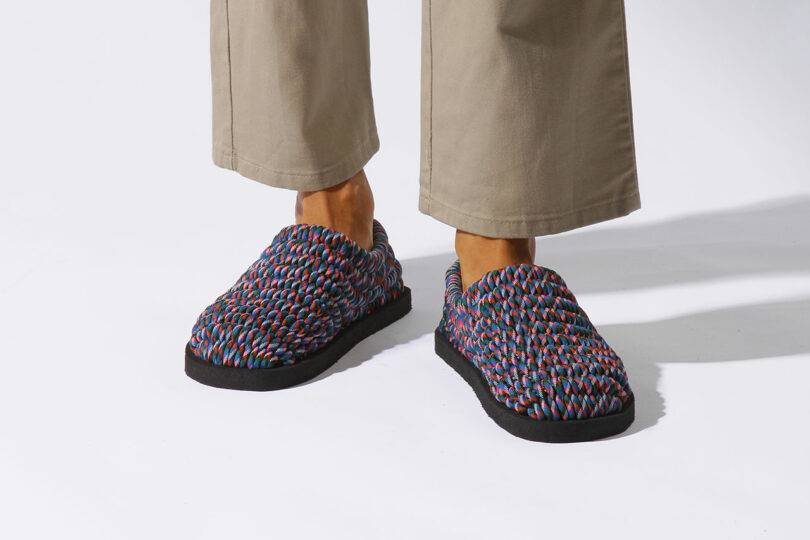 Person wearing beige pants and multicolored woven slip-on slippers with black soles, standing on a white surface.