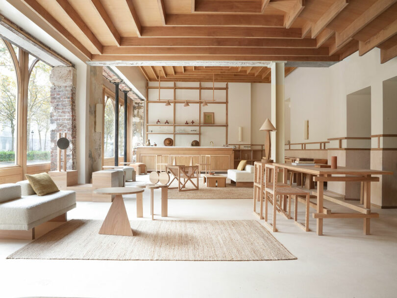 Spacious modern living room with wooden ceiling beams, minimalist light wood furniture, large windows, and neutral-toned decor.