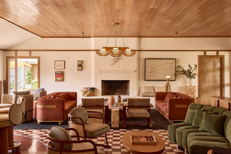 A spacious living room with a central white brick fireplace, various patterned armchairs and sofas, a wooden ceiling, light walls, and modern art on the walls.