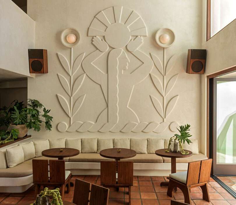 A sculpted wall mural of a woman with long hair holding a circle is centered above a beige cushioned bench, wooden tables, chairs, and potted plants in this sunlit room—perfect for your next Top 10 Travel 2025 inspiration spot.