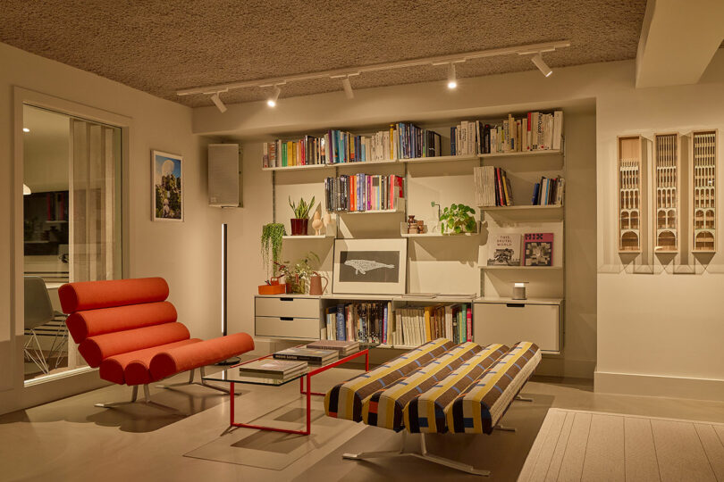 A modern living room with a red lounge chair, striped bench, glass coffee table, wall-mounted shelves filled with books, plants, framed art, and architectural models on the wall.