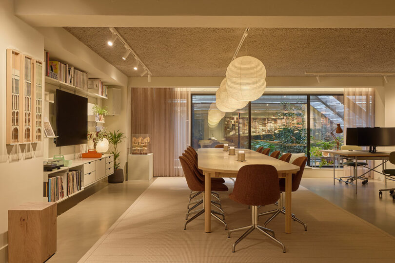 Modern office space with a long wooden table, brown chairs, hanging paper lantern lights, bookshelves, a TV, indoor plants, large windows, and a view of a green garden outside.
