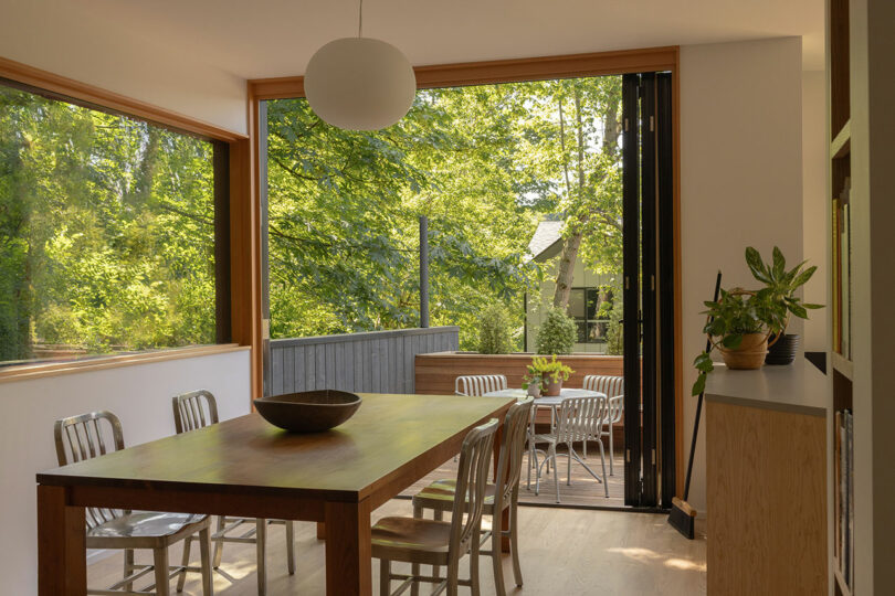 A modern dining room with wooden table and metal chairs opens to a deck with outdoor seating, surrounded by green trees and natural light.