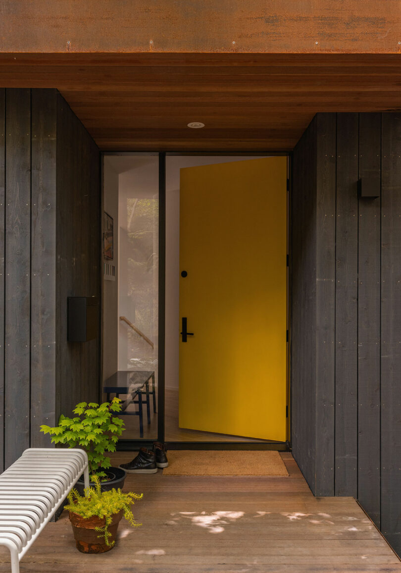 A modern entryway with dark wood siding, a partially open yellow door, a white bench, a potted plant, and shoes on a wooden porch.