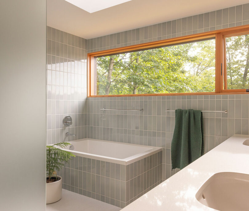 Modern bathroom with a built-in bathtub, light gray wall tiles, a large window with a view of green trees, and two green towels on a rack.
