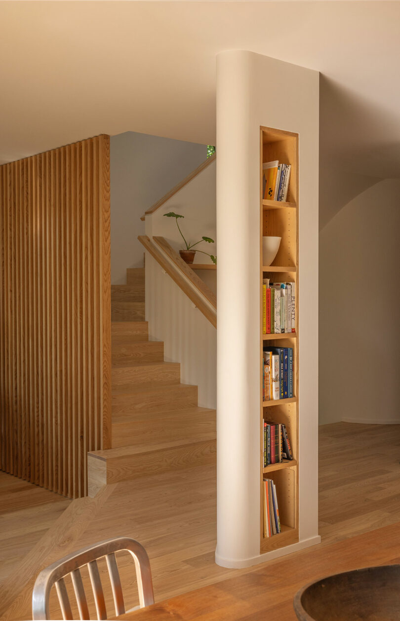 A built-in bookshelf is set into a column beside a wooden staircase with potted plants on the landing; wood tones dominate the minimalist, modern interior.