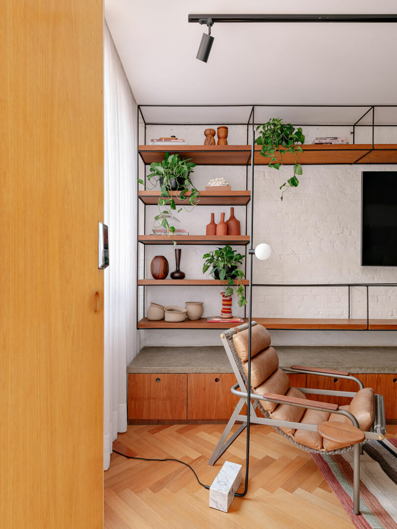 A modern living room corner with a tan leather chair, wood and metal shelving, potted plants, vases, a TV, and light wood floors.