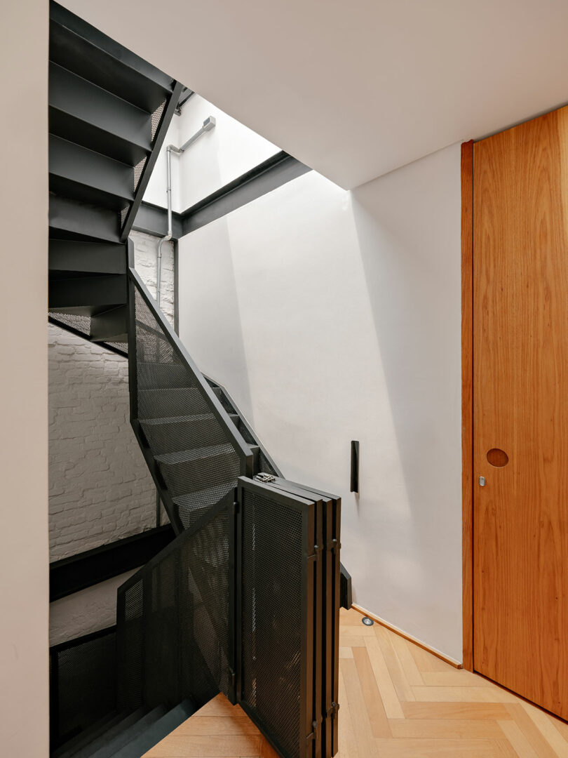Modern interior staircase with black metal steps and railing, adjacent to a white wall and wooden door, illuminated by natural light from above.