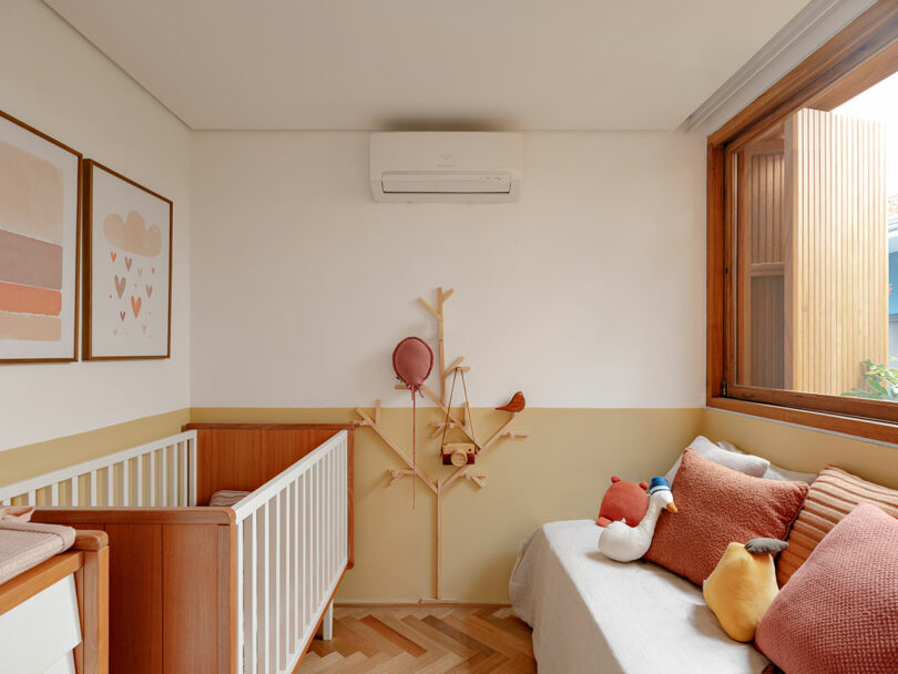 A cozy nursery with a crib, sofa with decorative pillows, wall art, a tree-shaped wall shelf, and an open window letting in natural light.