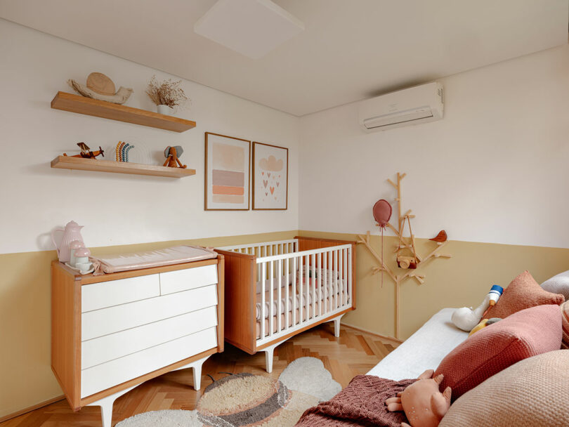 A neatly organized nursery features a crib, changing table, wall art, floating shelves, a sofa, and neutral-toned decor with pink and beige accents.