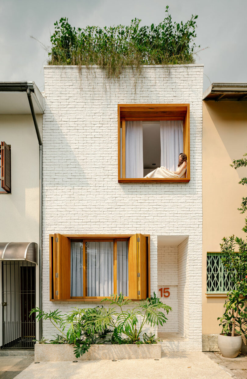 A modern white brick townhouse with large wooden-framed windows, plants on the roof, and a person sitting in the open upper window. The number 15 is visible near the entrance.